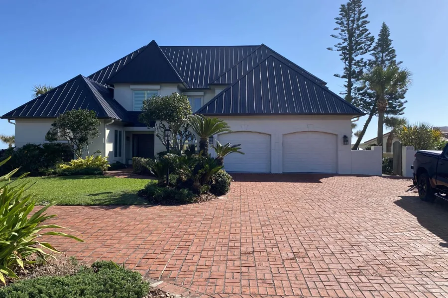 Two-story coastal home with a dark metal roof, white exterior walls, palm trees, and a brick paver driveway, the type of weather-exposed home A Classic Touch Painting repaints in Daytona Beach Shores, FL.